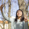Carefree woman grimacing and taking selfie photo outdoors.  Smiling pretty young lady posing, showing victory sign and using smartphone with trees in background. Selfie photograph concept.